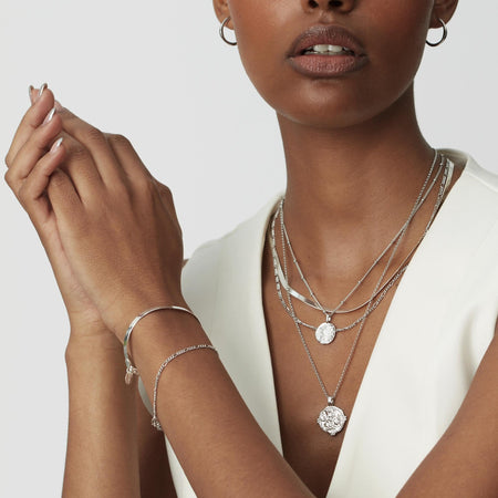 close up of model's neck wearing silver figaro chain necklace layered with a silver coin necklace on a bead chain, a silver snake chain necklace and a silver durga coin necklace with belcher chain.